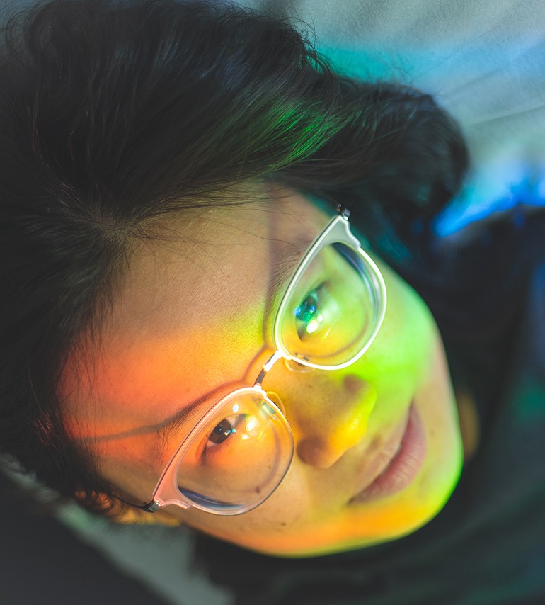 
Black hair swirling upwards, a face awash in gradients of the rainbow peers at the viewer with a slight smile, framed by her large, clear glasses and light blue bed sheets.